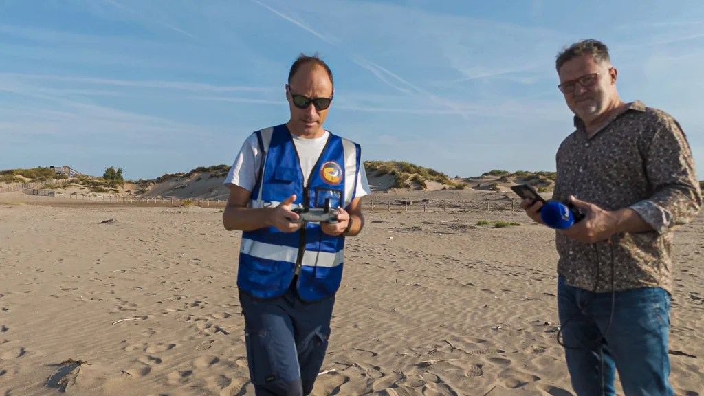 Deux hommes sur une plage bordée de dunes, l’un en gilet bleu tenant une télécommande